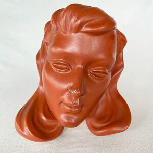 May include: A terracotta sculpture of a woman's head with closed eyes and a serene expression. The sculpture is a simple, minimalist design with a smooth, matte finish.