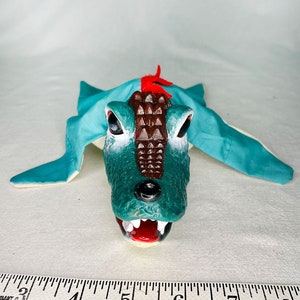 May include: A green and brown alligator puppet with a red crest and a white mouth. The puppet is lying on a white surface with a ruler in the foreground.