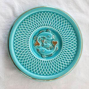 May include: A turquoise ceramic plate with a gold rim and a fish design. The plate features a raised fish motif in the center, surrounded by a lattice pattern. The fish are a bronze color. The plate is round and has a glossy finish.