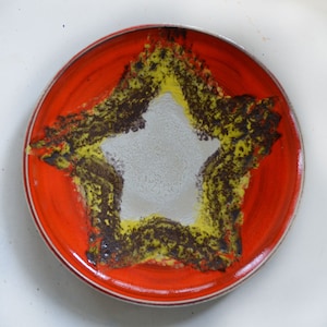 Puede incluir: Un plato de cerámica rojo con una estrella blanca en el centro. La estrella está rodeada por un diseño marrón y amarillo.