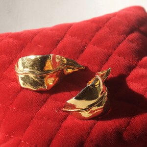 May include: A pair of gold-toned leaf-shaped earrings. The earrings are made of a textured metal and have a slightly curved shape.