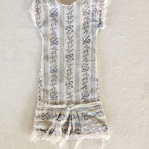 May include: A white cotton dress with a blue floral print and a ruffled hem. The dress has a simple, sleeveless design and is in a vintage style.