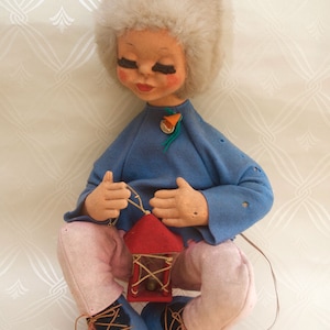Peut inclure: Une poupée en peluche portant une chemise bleue, un pantalon rose et un chapeau en fourrure blanche. La poupée tient une lanterne rouge avec une bordure dorée. La poupée a de longs cils et une expression douce.