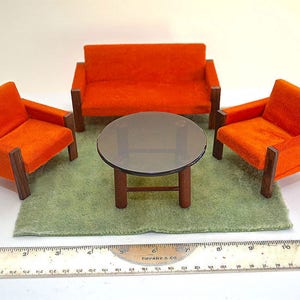 Op de afbeelding: Een set van drie oranje fluwelen stoelen en een ronde tafel met een glazen blad en een houten frame. De stoelen en de tafel staan op een groen tapijt.