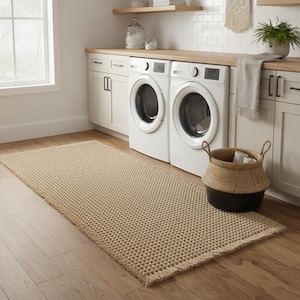Könnte beinhalten: Ein rechteckiger, beigefarbener Webteppich mit strukturiertem Muster und Fransen. Der Teppich liegt auf einem Holzboden in einem Waschraum, neben weißen Waschmaschinen und einem geflochtenen Korb.