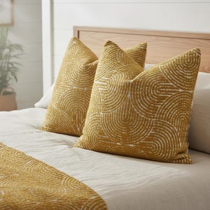 May include: Two mustard-yellow throw pillows with a white, semi-circular pattern rest on a bed with a matching bed runner. The pillows and runner have a textured appearance. The bed has a light-colored wooden headboard.