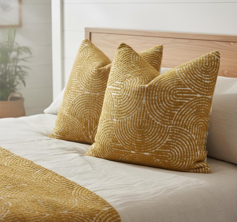 May include: Two mustard-yellow throw pillows with a white, semi-circular pattern rest on a bed with a matching bed runner. The pillows and runner have a textured appearance. The bed has a light-colored wooden headboard.
