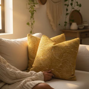 May include: Two mustard-yellow throw pillows with a white, concentric circle pattern rest on a cream-colored sofa. The pillows are square and textured, adding a decorative element to the living room. A person wearing a cream-colored sweater is seated on the sofa.