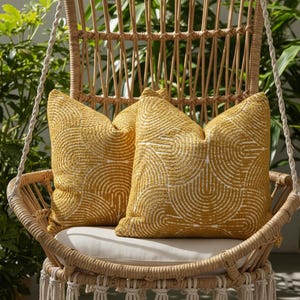 May include: Two yellow throw pillows with a white semi-circle pattern rest on a hanging wicker chair. The chair has a cream-colored cushion and macrame detailing. Green foliage is visible in the background.