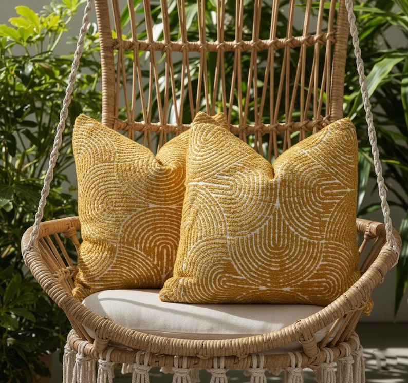 May include: Two yellow throw pillows with a white semi-circle pattern rest on a hanging wicker chair. The chair has a cream-colored cushion and macrame detailing. Green foliage is visible in the background.
