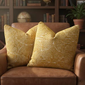 May include: Two mustard-yellow throw pillows with a white, semi-circular pattern rest on a brown leather armchair. The pillows have a textured appearance and are arranged side-by-side. The background includes a bookshelf, globe, and potted plant.