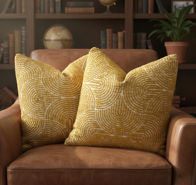 May include: Two mustard-yellow throw pillows with a white, semi-circular pattern rest on a brown leather armchair. The pillows have a textured appearance and are arranged side-by-side. The background includes a bookshelf, globe, and potted plant.