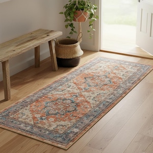 May include: A rectangular rug with a distressed, vintage design. The rug features a central pattern in orange and blue hues, framed by a blue border. It's placed on a wooden floor, adding warmth to the room. The rug's dimensions are in centimetres.