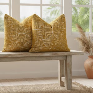 May include: Two mustard-yellow throw pillows with a white, semi-circular pattern. The pillows are placed on a light-colored wooden bench. The pillows add a decorative touch to any living space.
