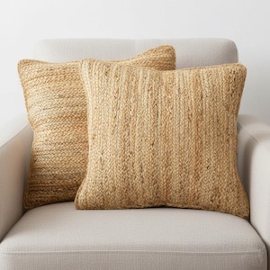 May include: Two square, woven jute pillows in shades of tan and beige rest on a light-colored armchair. The pillows have a textured, natural fiber appearance, adding a rustic touch to the decor. The pillows are approximately 18 inches square.