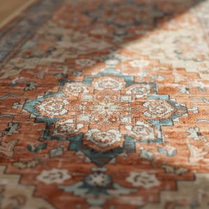 Peut inclure: Gros plan sur un tapis &agrave; motifs avec un motif de m&eacute;daillon central. Le tapis pr&eacute;sente une palette de couleurs chaudes d'orange rouille, de sarcelle et de cr&egrave;me, avec des motifs floraux et g&eacute;om&eacute;triques complexes. La texture du tapis est visible, sugg&eacute;rant une construction tiss&eacute;e.