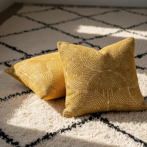 May include: Two mustard-yellow throw pillows with a white, semi-circular pattern. The pillows are square-shaped and rest on a cream-colored rug with a black geometric design. The pillows have a textured appearance.