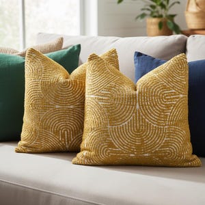 May include: Two mustard-yellow throw pillows with a white, semi-circular pattern. The pillows are on a light gray couch, with other pillows in shades of green and blue. The pillows have a textured appearance and add a decorative touch to the living room.