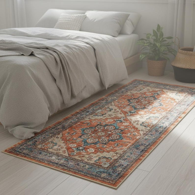 Peut inclure: Un tapis rectangulaire avec un motif orn&eacute; d'inspiration vintage. Le tapis pr&eacute;sente un m&eacute;daillon central dans des tons d'orange, de bleu et de cr&egrave;me, encadr&eacute; d'une bordure aux couleurs similaires. Il est pos&eacute; sur un sol en bois clair pr&egrave;s d'un lit.