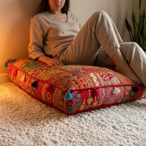 May include: A large, square floor cushion with a vibrant red background and intricate gold embroidery. The cushion features colorful patches, tassels, and a decorative border. It is placed on a white rug.
