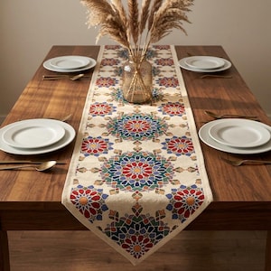 May include: A rectangular table runner featuring a repeating geometric mandala pattern in shades of red, blue, green, and yellow on a cream-colored fabric. The runner is centered on a wooden dining table with four white plates and gold cutlery.