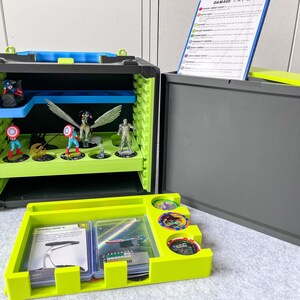 May include: A black and green storage case with a blue handle and accents. The case is open, revealing shelves with superhero figurines. A yellow tray holds cards and tokens. A blue and white card is visible.