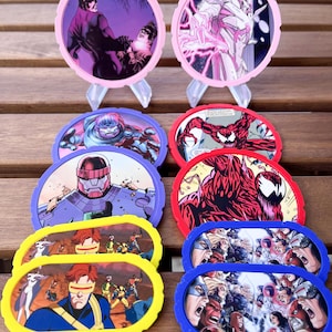 Puede incluir: Una colección de posavasos coloridos, de forma circular y ovalada, con personajes de cómics. Los posavasos tienen bordes de varios colores, incluyendo rosa, morado, rojo, amarillo y azul. Las imágenes en los posavasos representan escenas de acción y retratos de personajes.