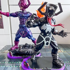 Peut inclure: Deux figurines articulées sur des socles violets. L'une est une figurine de Galactus violette et bleu métallique. L'autre est un hybride Venom-Galactus noir avec un emblème d'araignée blanc, une longue langue rouge et un grand casque avec des flammes orange.