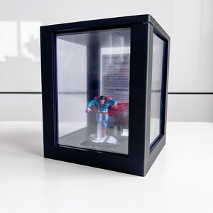 Puede incluir: Una vitrina negra con paneles de vidrio transparente alberga una figura de Superman. La figura, en pose de vuelo, lleva un traje azul, una capa roja y botas. La vitrina está sobre una superficie blanca.