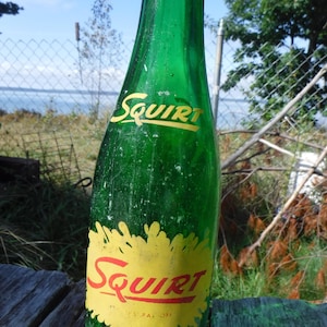 May include: A vintage green glass Squirt soda bottle. The bottle has a yellow and red label with the word "Squirt" in a stylized font. The bottle is sitting on a wooden surface, with a natural outdoor background.