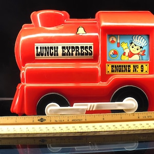 May include: A red plastic toy train lunchbox with the words "LUNCH EXPRESS" and "ENGINE NO. 9" printed on it. The train has black wheels, a white bar, and a cartoon image of a conductor. A ruler is placed in front of the train.