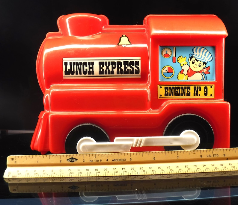 May include: A red plastic toy train lunchbox with the words "LUNCH EXPRESS" and "ENGINE NO. 9" printed on it. The train has black wheels, a white bar, and a cartoon image of a conductor. A ruler is placed in front of the train.
