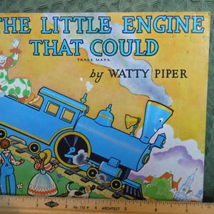 Könnte beinhalten: Ein Vintage-Kinderbuchcover mit dem Titel "THE LITTLE ENGINE THAT COULD" von Watty Piper. Das Cover zeigt einen blauen Zeichentrickzug, einen Clown und andere Charaktere auf gelbem Hintergrund. Das Buch ist etwa 25 cm breit.