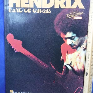 Könnte beinhalten: Ein Musikbuch mit dem Titel "Jimi Hendrix Band of Gypsys Transcribed" mit dunklem Hintergrund. Das Cover zeigt ein Porträt von Jimi Hendrix, der eine rote Gitarre spielt. Das Hal Leonard-Logo befindet sich unten.