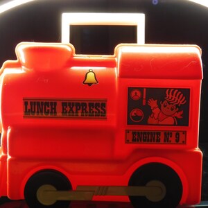 May include: A bright red plastic lunchbox shaped like a train. The lunchbox has black wheels, a handle, and the words "LUNCH EXPRESS" printed on the side. It also features a cartoon image and the text "ENGINE No. 9".