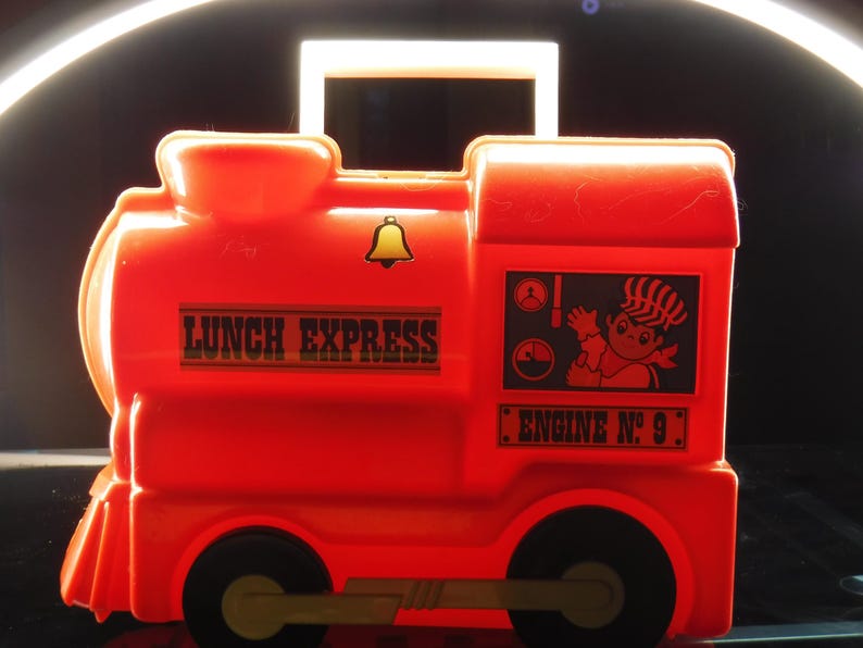 May include: A bright red plastic lunchbox shaped like a train. The lunchbox has black wheels, a handle, and the words "LUNCH EXPRESS" printed on the side. It also features a cartoon image and the text "ENGINE No. 9".