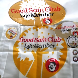 Könnte beinhalten: Eine Sammlung von Good Sam Club Life Member-Artikeln. Enthält ein ovales Schild, eine Flagge, einen Aufkleber, zwei Anstecknadeln und einen runden Aufkleber, alle mit dem Logo des Clubs und Text in Rot und Schwarz auf gelbem und weißem Hintergrund.