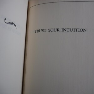 K&ouml;nnte beinhalten: Offene Buchseiten mit dem Text "TRUST YOUR INTUITION" in einer Serifenschrift. Eine dekorative schwarze Verzierung befindet sich in der oberen linken Ecke. Die Seiten sind cremefarben mit einer leichten Textur.