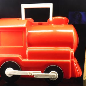 May include: A bright red plastic toy train with black wheels and a white handle. The toy train has a rectangular window and a rounded top. The wheels are black with white accents. The handle is white and rectangular.