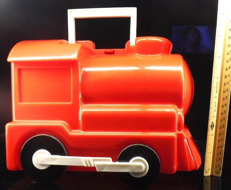 May include: A bright red plastic toy train with black wheels and a white handle. The toy train has a rectangular window and a rounded top. The wheels are black with white accents. The handle is white and rectangular.