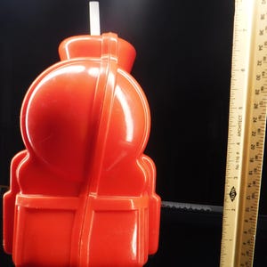 May include: A bright orange plastic container with a white straw. The container has a rounded top and a rectangular base. A ruler is next to the container, showing measurements in inches.