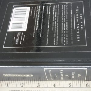 K&ouml;nnte beinhalten: Ein schwarzes Hardcover-Buch mit dem Titel "The Book of Answers" mit wei&szlig;em Text. Das Buch hat eine rechteckige Form und scheint geschlossen zu sein. Der Buchr&uuml;cken tr&auml;gt den Titel und den Namen des Autors. Das Buch liegt auf einer Oberfl&auml;che.