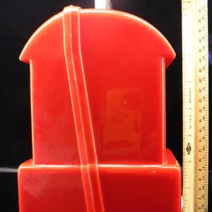 May include: A bright red plastic house-shaped container with a white rectangular top. A ruler is placed next to the container. The container has a rectangular base and a curved roof. A red strap runs down the front.