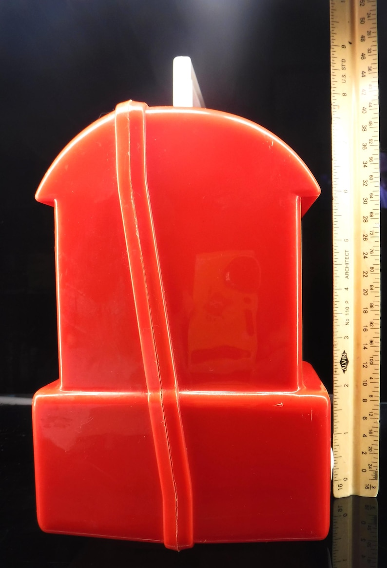 May include: A bright red plastic house-shaped container with a white rectangular top. A ruler is placed next to the container. The container has a rectangular base and a curved roof. A red strap runs down the front.