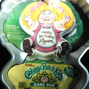 Puede incluir: Molde para pastel Cabbage Patch Kids plateado con un diseño de dibujos animados. El molde incluye una etiqueta amarilla y verde con las palabras "Cabbage Patch Kids CAKE PAN". El diseño del pastel dice "Happy Birthday Lauren".