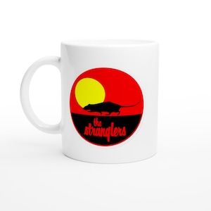 Puede incluir: Taza de cerámica blanca con un diseño. El diseño presenta un círculo rojo con un sol amarillo, una silueta negra de rata y el texto "the stranglers" en blanco, con una fuente estilizada.