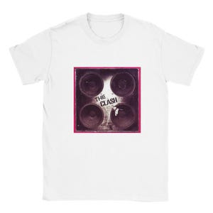 Puede incluir: Camiseta blanca con un gráfico de cuatro conos de altavoz en un marco cuadrado con un borde rosa. El texto "THE CLASH" está impreso en blanco en el gráfico. La camiseta está hecha de un material suave y cómodo.