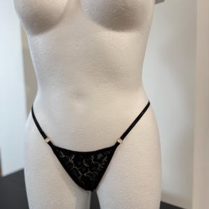 Könnte beinhalten: Schwarzer Spitzen-String mit zartem Blumenmuster. Der String hat dünne Träger und kleine goldfarbene Ringe an den Seiten. Die Unterwäsche wird auf einer weißen Schaufensterpuppe präsentiert.