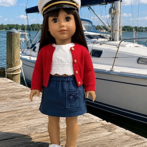 May include: A doll with long brown hair wearing a white sailor hat with a gold anchor emblem, a red knit cardigan with gold buttons, a white cropped top with lace trim, a navy blue skirt with a small pocket and bow, and white glittery slip-on shoes.