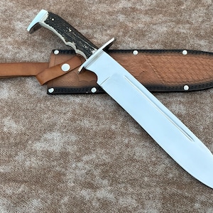 May include: A large, polished steel hunting knife with a brown and black antler handle and a leather sheath. The knife has a wide blade and a full tang construction. The sheath is brown leather with a belt loop.